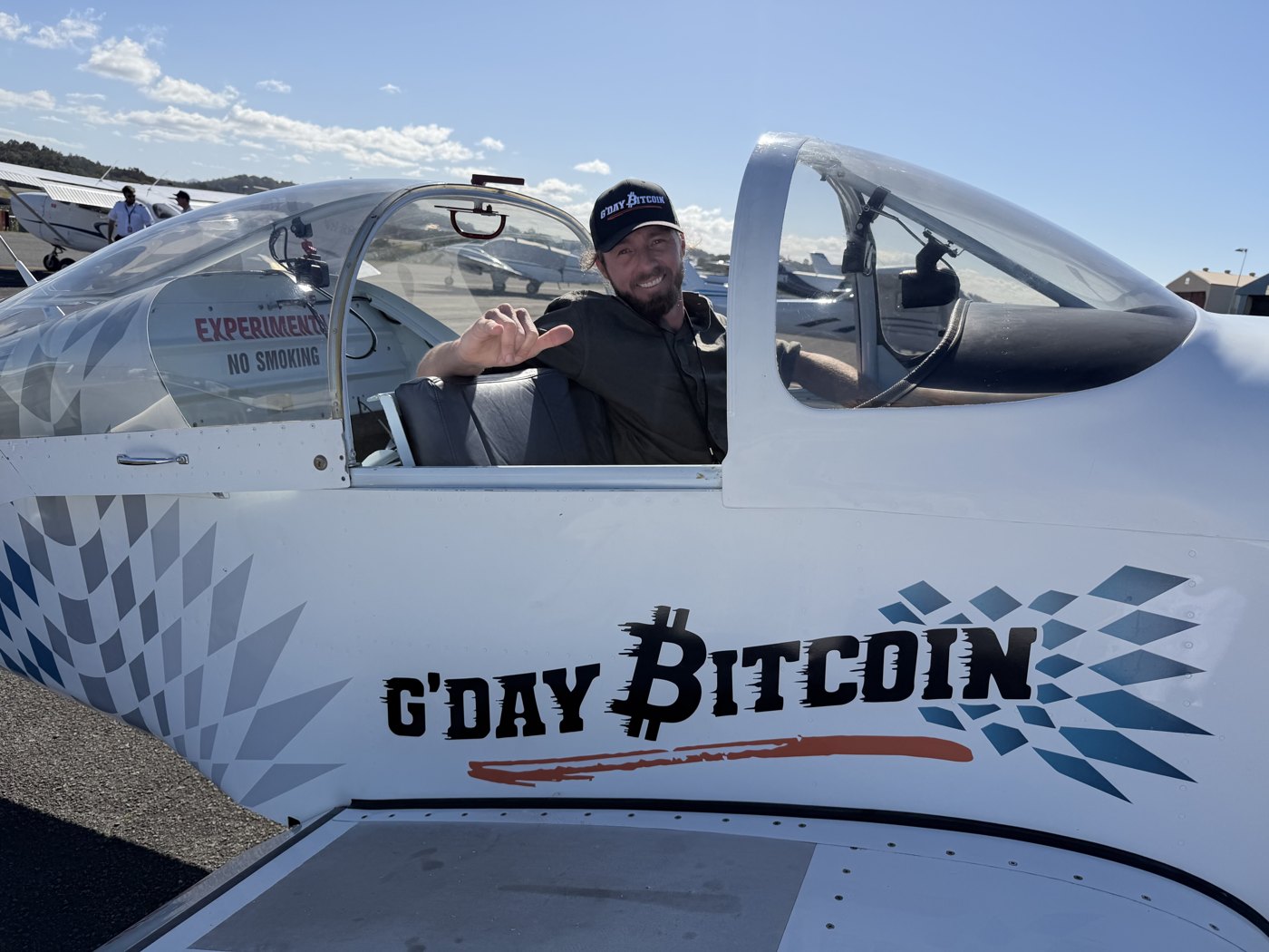 A smiling man wearing a G’day Bitcoin cap in the cockpit of a G’day Bitcoin-branded aircraft